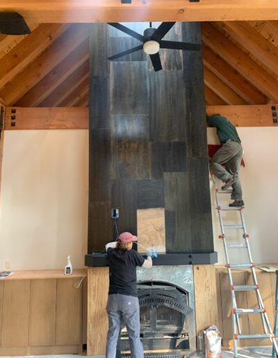 Two individuals working on the installation of a wall feature above a fireplace in a room with exposed wooden beams.