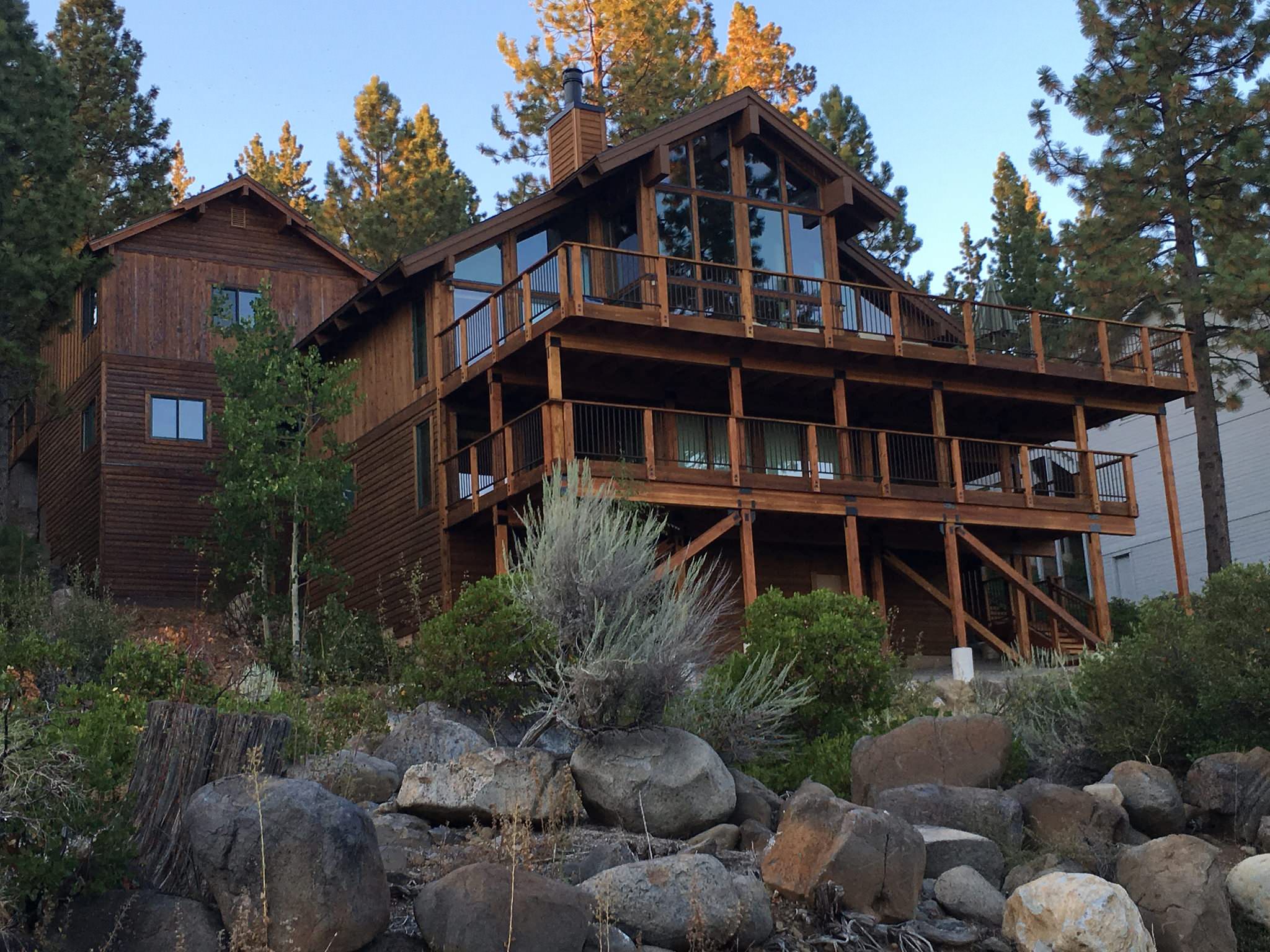 A multi-story wooden cabin with expansive decks located on a rocky hillside with trees in the background during dusk.