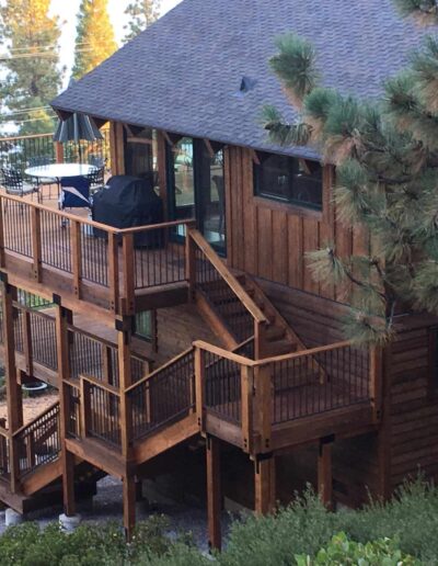 A wooden cabin with a large deck surrounded by pine trees during the evening.